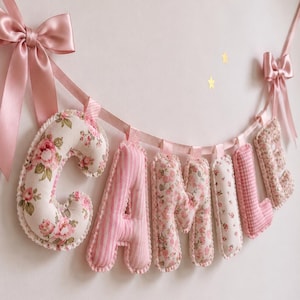 May include: A decorative fabric banner spelling the name CAMILLE with plush, stuffed letters. The letters feature alternating pink floral, striped, and gingham patterns, suspended by a pink satin ribbon with bows at each end of the string.