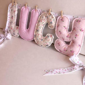 May include: Four stuffed fabric letters spelling LUCY, each featuring distinct floral or gingham patterns in shades of pink and cream. The letters are suspended from a white ribbon with a floral print using small wooden clothespins.