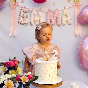 May include: A birthday cake with a white floral pattern, topped with a small plush rabbit and a white number two candle. The cake sits on a wooden stand, surrounded by pink and white flowers, with a banner spelling EMMA in the background.