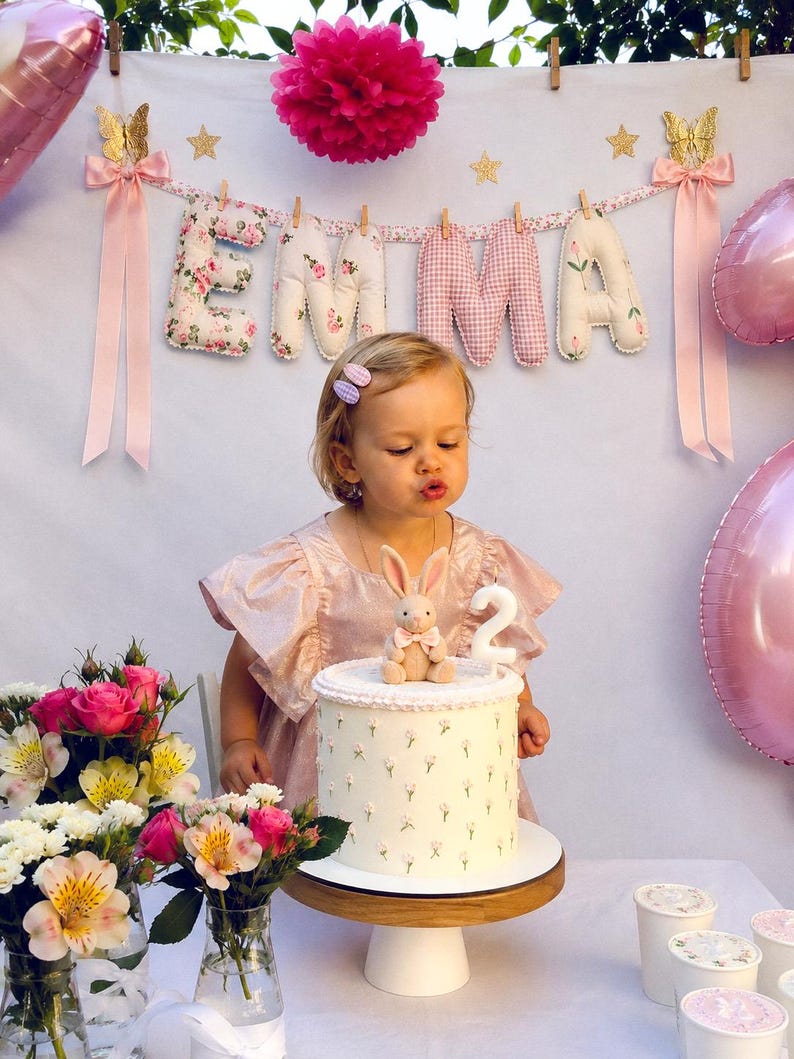 May include: A birthday cake with a white floral pattern, topped with a small plush rabbit and a white number two candle. The cake sits on a wooden stand, surrounded by pink and white flowers, with a banner spelling EMMA in the background.