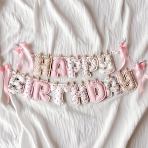 May include: A fabric banner spelling out HAPPY BIRTHDAY with stuffed, individual letters in various pink, white, and floral patterns. The letters are clipped to a string with small wooden clothespins and decorated with light pink satin ribbons.