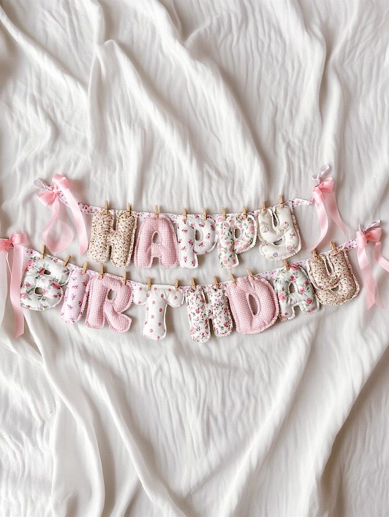 May include: A fabric banner spelling out HAPPY BIRTHDAY with stuffed, individual letters in various pink, white, and floral patterns. The letters are clipped to a string with small wooden clothespins and decorated with light pink satin ribbons.