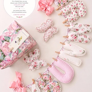 May include: A fabric banner spelling the word SUNSHINE with stuffed letters attached to a ribbon by small wooden clothespins. The letters feature floral and polka dot patterns in pink, white, and green. Two fabric butterflies and a gift box are included.