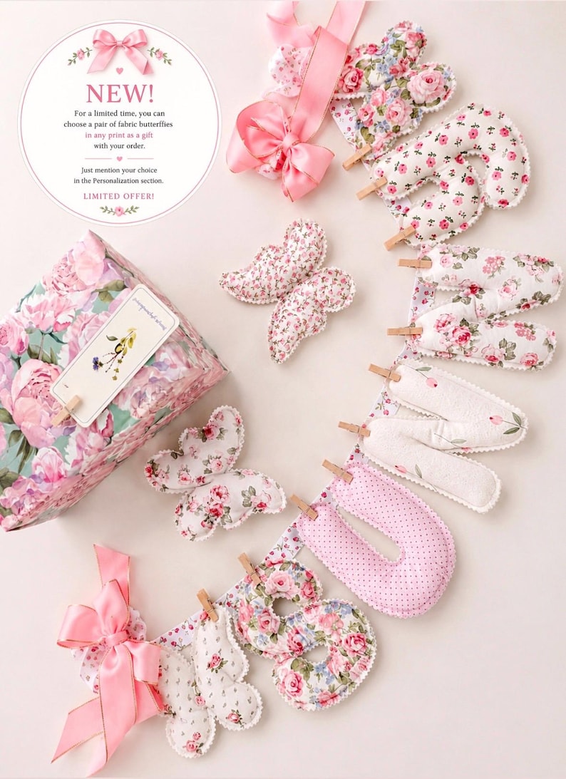 May include: A fabric banner spelling the word SUNSHINE with stuffed letters attached to a ribbon by small wooden clothespins. The letters feature floral and polka dot patterns in pink, white, and green. Two fabric butterflies and a gift box are included.