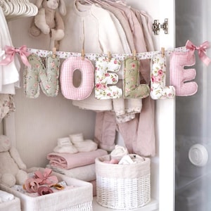 May include: A fabric banner spelling the name NOELLE with stuffed letters hanging from a floral ribbon. The letters alternate between a sage green floral print, a pink and white gingham check, and a white fabric with a pink rose floral pattern.