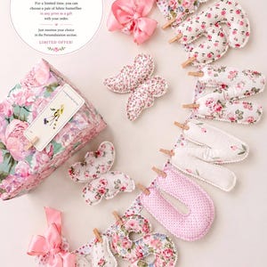 May include: A fabric banner spelling the word SUN with stuffed letters attached to a string by small wooden clips, featuring pink floral and polka dot patterns. Includes two fabric butterflies, pink satin bows, and a floral gift box.