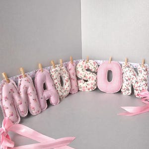 May include: A decorative banner spelling the name MADISON with individual stuffed fabric letters. The letters feature alternating pink floral patterns and a pink gingham check print, each attached to a white ribbon with small wooden clothespins.