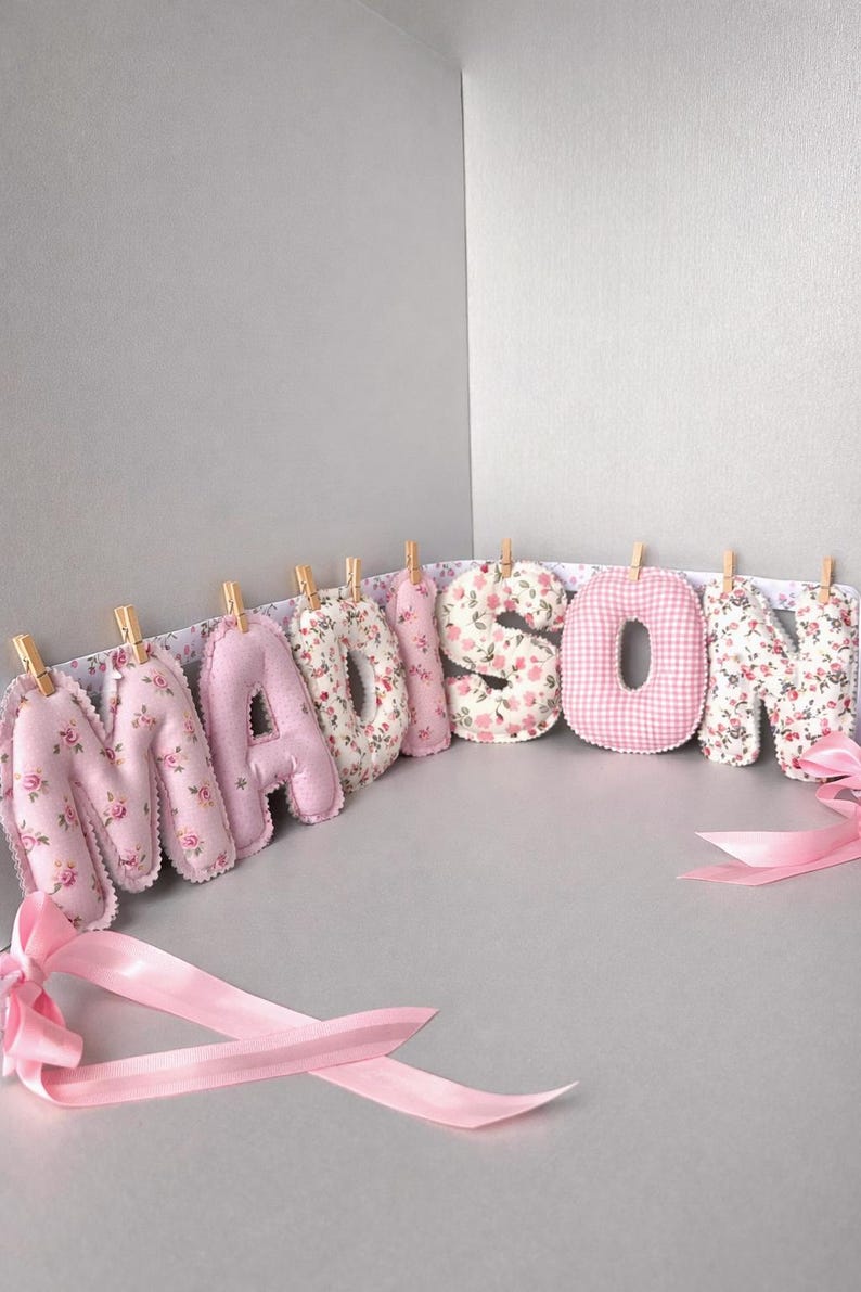May include: A decorative banner spelling the name MADISON with individual stuffed fabric letters. The letters feature alternating pink floral patterns and a pink gingham check print, each attached to a white ribbon with small wooden clothespins.