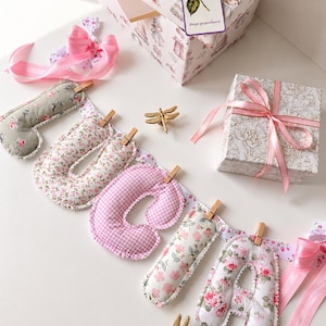 May include: A fabric banner spelling the name LUCIA with stuffed, padded letters in floral and pink gingham patterns, attached to a ribbon with wooden clips. Includes two gold-toned dragonfly ornaments and two gift boxes with pink satin ribbons.
