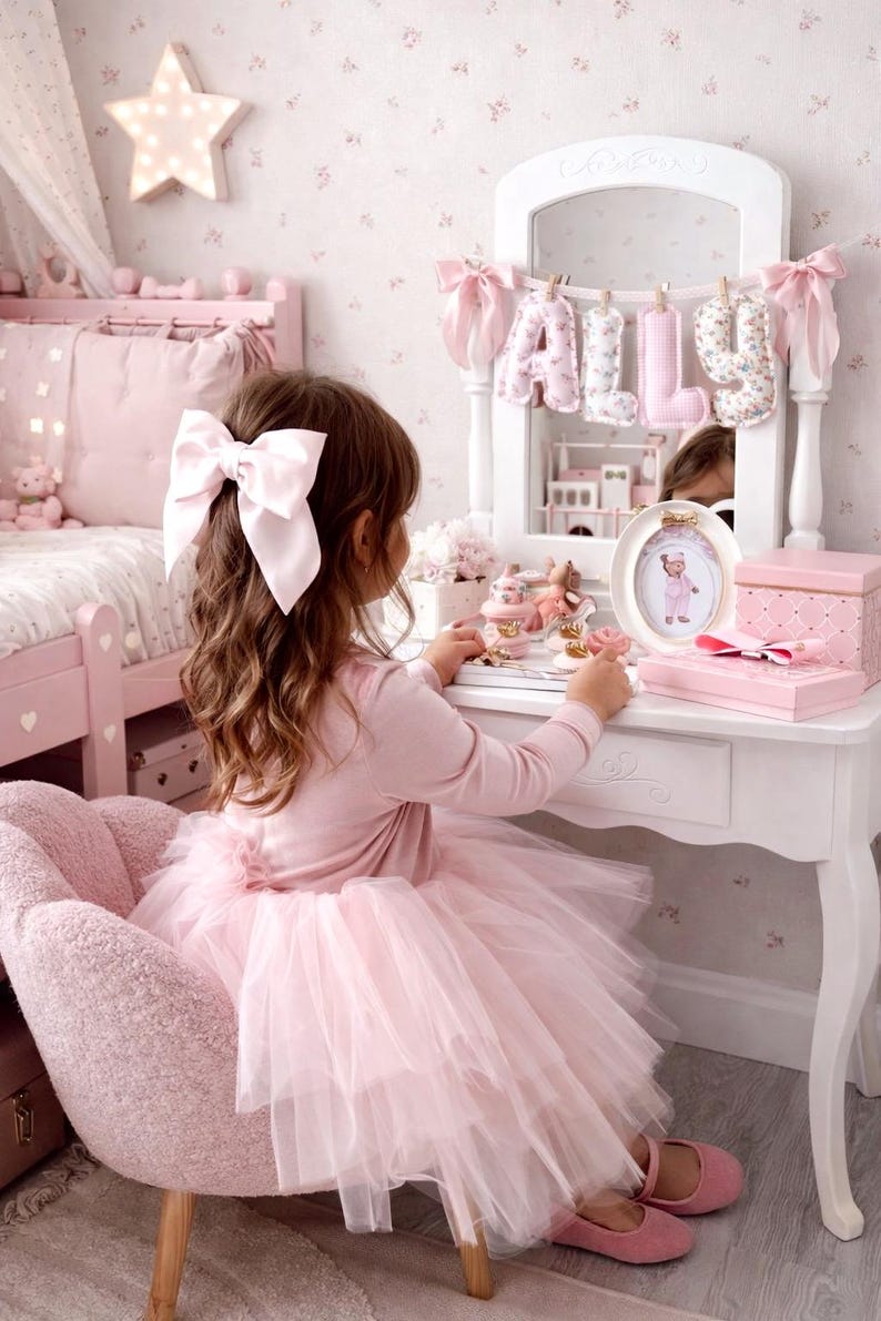 May include: A white vanity table with a mirror featuring a hanging banner that spells out the name ALLY in fabric letters. The vanity is decorated with a small oval framed picture, a pink box, and various small trinkets on the tabletop surface.