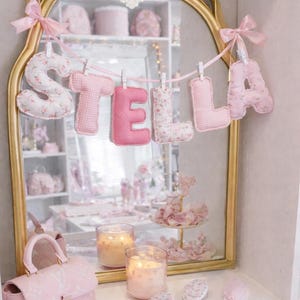 May include: A gold-framed mirror with a bow top features a hanging banner of plush fabric letters spelling STELLA. The letters alternate between pink gingham and a white floral print, suspended by small white clips on a pink ribbon.
