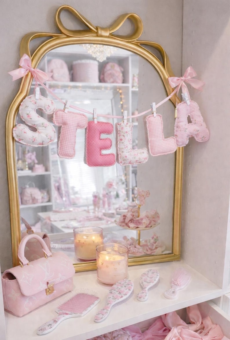May include: A gold-framed mirror with a bow top features a hanging banner of plush fabric letters spelling STELLA. The letters alternate between pink gingham and a white floral print, suspended by small white clips on a pink ribbon.