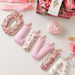 May include: A fabric banner spelling the name OLIVIA with stuffed letters attached to a ribbon by wooden clips. The letters feature alternating pink gingham and floral patterns. Includes a pink heart-shaped box, pink ribbons, and a floral gift bag.