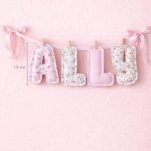 May include: A fabric banner spelling the name ALLY with four stuffed letters hanging from a ribbon with wooden clips and pink satin bows at each end. Each letter measures 15 cm in height and features a mix of floral and gingham patterns.
