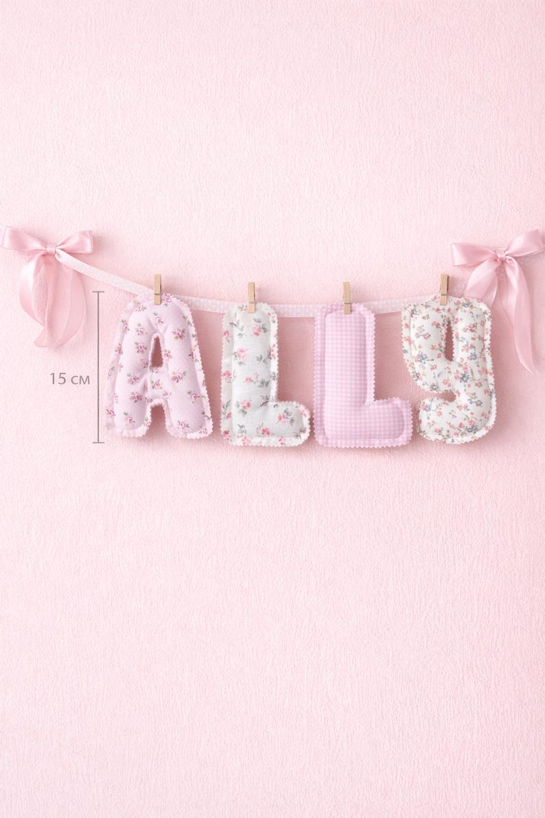 May include: A fabric banner spelling the name ALLY with four stuffed letters hanging from a ribbon with wooden clips and pink satin bows at each end. Each letter measures 15 cm in height and features a mix of floral and gingham patterns.