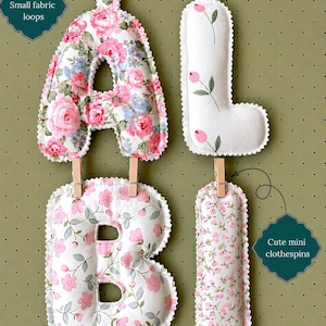 May include: Four stuffed fabric letters spelling ALBI, featuring floral patterns in pink, white, and green. The letters have small fabric loops at the top and are connected by wooden mini clothespins. Text includes Small fabric loops, Cute mini clothespins, and Please choose your preferred hanging option!