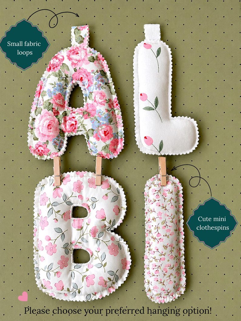 May include: Four stuffed fabric letters spelling ALBI, featuring floral patterns in pink, white, and green. The letters have small fabric loops at the top and are connected by wooden mini clothespins. Text includes Small fabric loops, Cute mini clothespins, and Please choose your preferred hanging option!