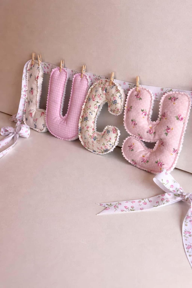 May include: Four stuffed fabric letters spelling LUCY, each featuring distinct floral or gingham patterns in shades of pink and cream. The letters are suspended from a white ribbon with a floral print using small wooden clothespins.