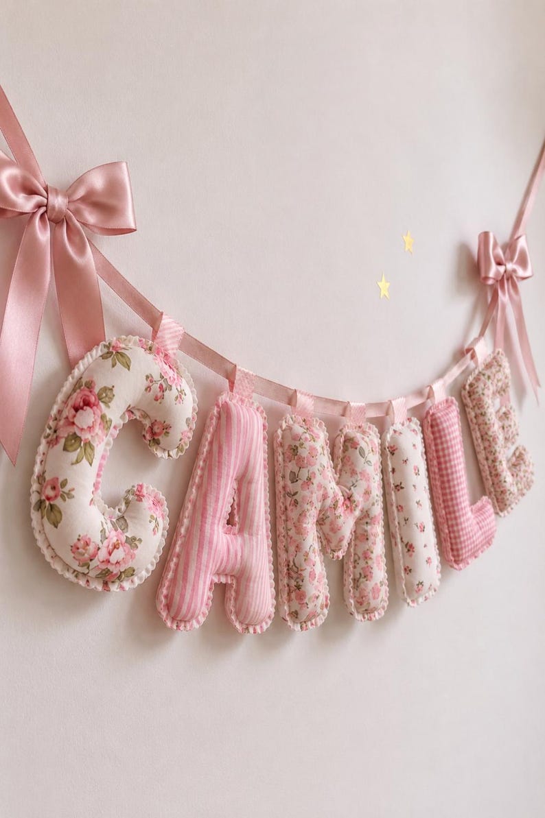 May include: A decorative fabric banner spelling the name CAMILLE with plush, stuffed letters. The letters feature alternating pink floral, striped, and gingham patterns, suspended by a pink satin ribbon with bows at each end of the string.