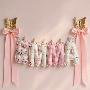 May include: A decorative banner spelling the name EMMA with stuffed fabric letters attached to a floral ribbon by wooden clips. The letters feature pink floral and gingham patterns. Each end is adorned with a gold butterfly and a pink satin bow.
