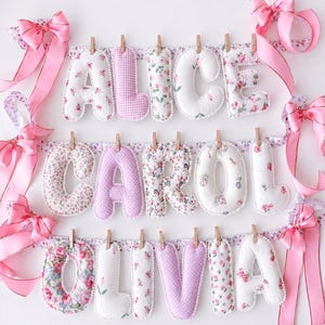 May include: A decorative banner featuring three rows of stuffed fabric letters spelling ALICE, CAROL, and OLIVIA. The letters are made from white and pink floral and gingham patterned fabric, suspended by wooden clips on ribbons with pink bows.