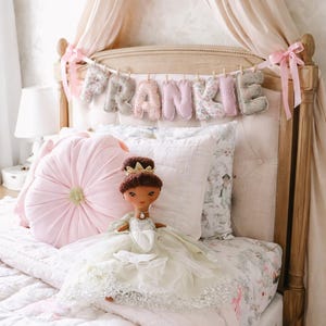 May include: A plush doll with dark brown hair and a gold crown, wearing a layered white tulle dress with silver accents. The doll sits on a bed with a pink tufted round pillow, a white quilted pillow, and a banner spelling FRANKIE in fabric letters.