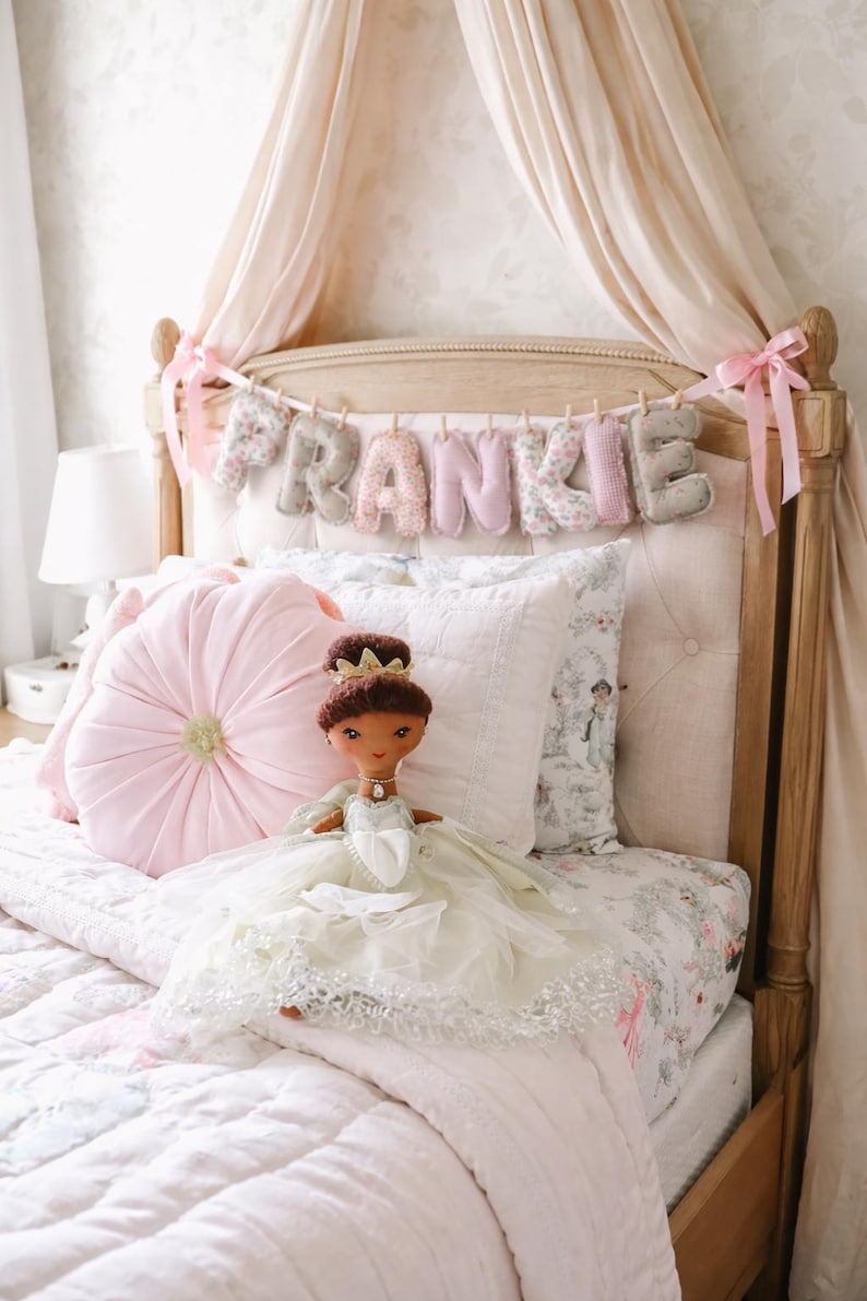 May include: A plush doll with dark brown hair and a gold crown, wearing a layered white tulle dress with silver accents. The doll sits on a bed with a pink tufted round pillow, a white quilted pillow, and a banner spelling FRANKIE in fabric letters.