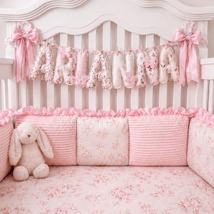 May include: A crib bedding set featuring a pink floral pattern, including a fitted sheet, ruffled bumper pads, and a plush bunny toy. A fabric banner spelling the name ARIANNA hangs from the headboard, accented with pink bows and butterflies.