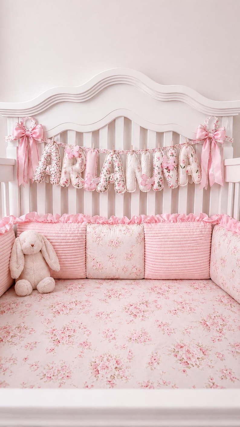 May include: A crib bedding set featuring a pink floral pattern, including a fitted sheet, ruffled bumper pads, and a plush bunny toy. A fabric banner spelling the name ARIANNA hangs from the headboard, accented with pink bows and butterflies.