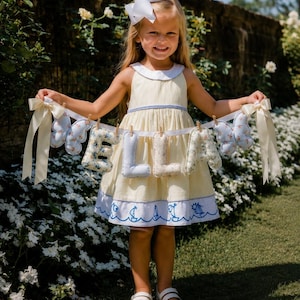 May include: A fabric banner featuring the name ELLA in stuffed, patterned letters, flanked by two light blue butterfly-shaped ornaments. The letters and butterflies are attached to a white ribbon with small wooden clips and cream-colored bows.