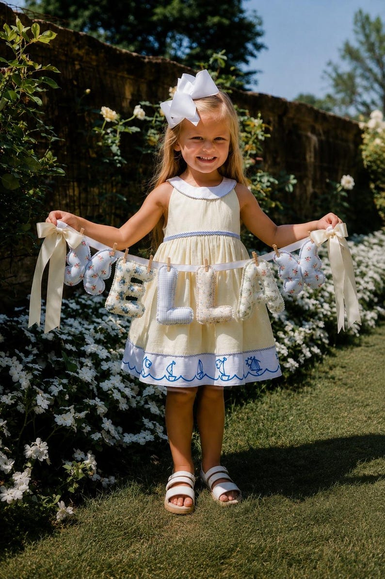 May include: A fabric banner featuring the name ELLA in stuffed, patterned letters, flanked by two light blue butterfly-shaped ornaments. The letters and butterflies are attached to a white ribbon with small wooden clips and cream-colored bows.