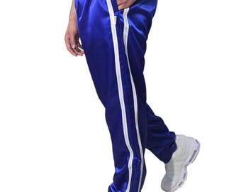 May include: Royal blue satin track pants with white side stripes. The pants have a drawstring waist and side pockets. The model is wearing white sneakers. The pants are a casual style.