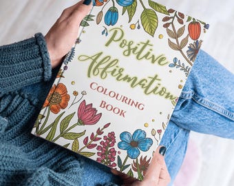 May include: A coloring book titled "Positive Affirmations Colouring Book" with floral illustrations in orange, pink, blue, and green. The book is held open, displaying the cover design.