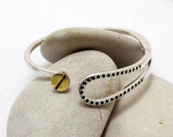 May include: A silver bangle bracelet with a black stone design and a gold accent. The bracelet is open and has a simple, modern design.