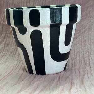 Può includere: Un vaso per piante conico caratterizzato da un motivo astratto dipinto a mano in bianco e nero. Il bordo è decorato con strisce verticali alternate, mentre il corpo presenta forme organiche curve in un design monocromatico ad alto contrasto.