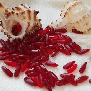 May include: A pile of red glass beads in the shape of teardrops, scattered on a white fabric background. Two seashells are visible in the background.