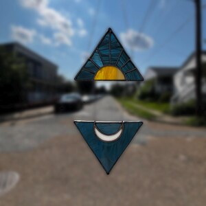 May include: Two stained glass suncatchers. The top one is a triangle with a yellow sunburst design and blue glass. The bottom one is a triangle with a white crescent moon and blue glass. Both have black metal borders.