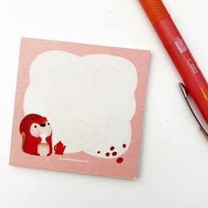 May include: Pink notepad with a red squirrel illustration holding a teacup. The squirrel is looking at a cloud-shaped note space with red coffee beans scattered below. The notepad has the text "thelittleredhouse.co" at the bottom.