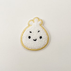 May include: A white, fuzzy, embroidered patch shaped like a dumpling with a smiling face. The patch has a gold outline.