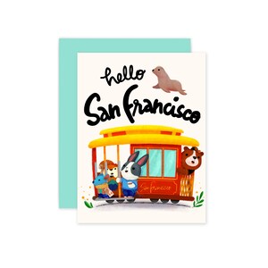 May include: Greeting card with a light teal envelope featuring a whimsical illustration. The card depicts a yellow cable car filled with animal characters, the text "hello San Francisco", and a seal. The card is on a white background.