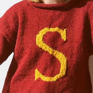 May include: A red knitted sweater with a large yellow letter "S" on the front. The sweater has a crew neck and long sleeves. The knitwear is a warm, cozy style.
