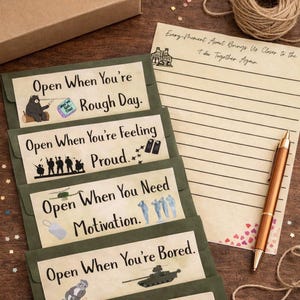 Puede incluir: Sobres verdes oliva con mensajes impresos como "Open When You're Rough Day" e ilustraciones. También hay un papel rayado con texto y un bolígrafo, junto con una madeja de hilo y una caja de cartón.