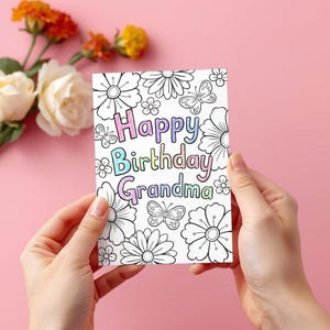 May include: A greeting card with the words "Happy Birthday Grandma" in colorful letters, surrounded by black and white floral and butterfly illustrations. The card is held against a pink background, with flowers in the upper left corner.