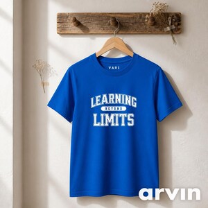 以下が含まれることがあります： 胸元にカレッジフォントで「LEARNING BEYOND LIMITS」という白い文字がプリントされた、ロイヤルブルーの半袖Tシャツ。内側の襟のラベルには「VAR1」という白い文字が表示されています。Tシャツは木製のハンガーに掛けられています。