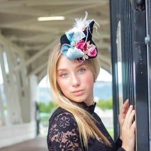 May include: A black fascinator hat with a small brim, adorned with light blue bows, pink roses, and white feathers. The hat is worn by a person wearing a black lace top.