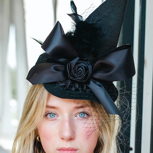 May include: A young person with blonde hair wearing a black witch hat with a black bow and a black rose. The hat has a black veil.