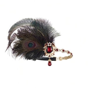 May include: A vintage-style headband featuring a large peacock feather and a decorative jewel-encrusted centerpiece. The centerpiece has a large red jewel and smaller red jewels. The headband is black and gold, with a dangling red jewel.
