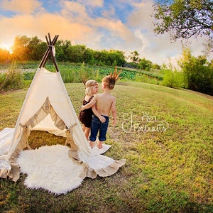 May include: Two children dressed in Native American costumes stand near a teepee. The girl is wearing a brown dress and a feather headdress. The boy is wearing a blue shirt and a feather headdress. They are both looking at each other.