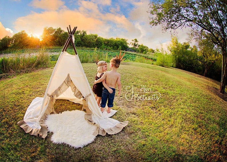 May include: Two children dressed in Native American costumes stand near a teepee. The girl is wearing a brown dress and a feather headdress. The boy is wearing a blue shirt and a feather headdress. They are both looking at each other.