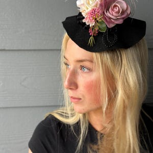 May include: A black witch hat with a wide brim, adorned with pink and white fabric flowers, green leaves, and pink feathers. The hat is decorated with a beaded accent. The hat is a festive accessory.